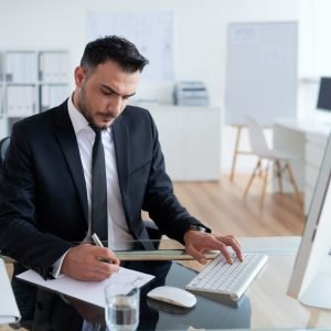 hombre-caucasico-en-traje-de-negocios-sentado-en-la-oficina-trabajando-en-la-computadora-y-escribiendo-en-papel